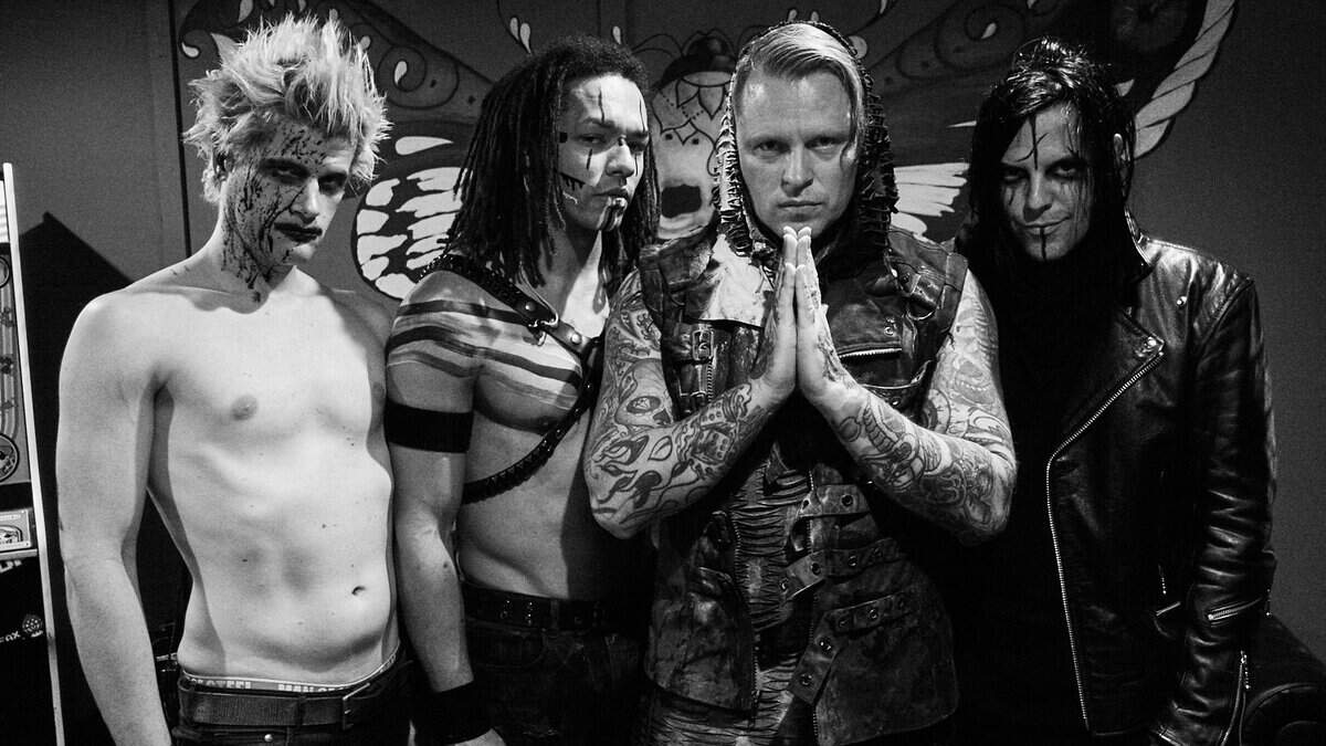 Four men in punk/goth outfits with face paint pose together, the central figure pressing his hands in a prayer-like gesture.
