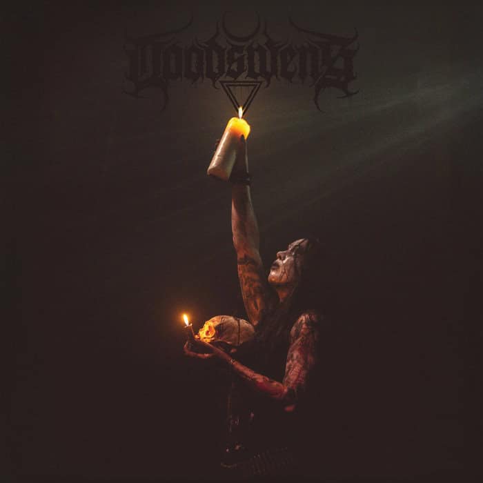 Dark stage performance: tattooed, face-painted musician raises a lit bottle-mounted flame while holding a candle and a skull prop nearby, with a band logo above.