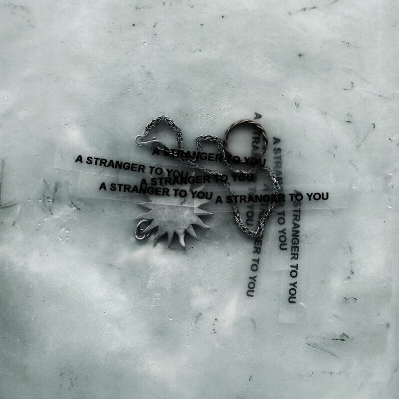 Necklace with a sun pendant on a weathered surface, wrapped by strips of tape reading 'A STRANGER TO YOU'.