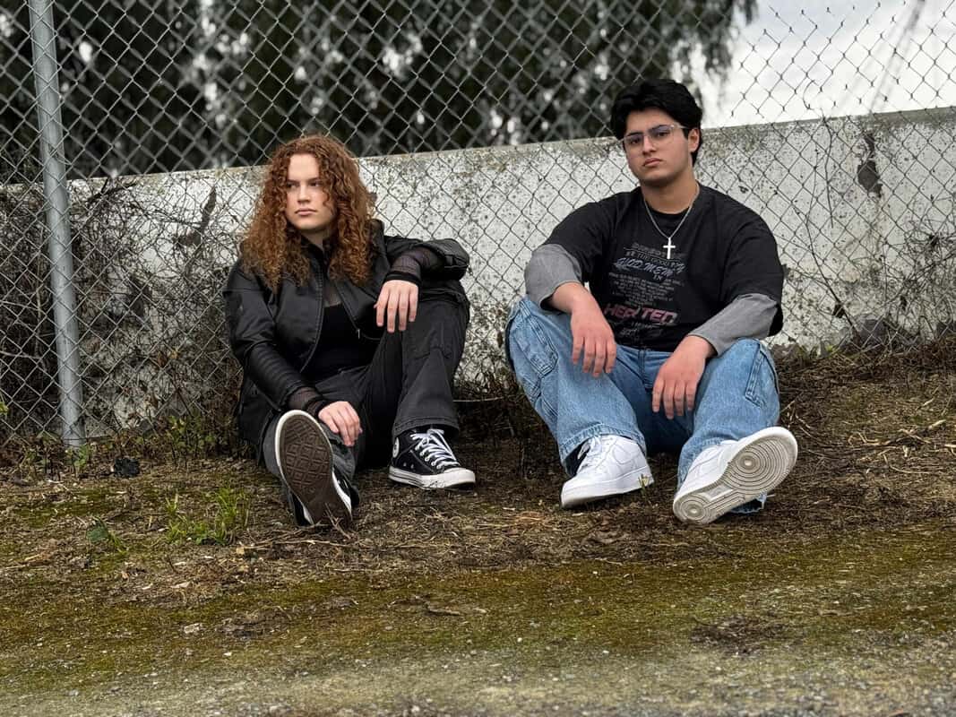 Two friends sit on the dirt ground against a chain-link fence; left: curly red-haired person in a black jacket and sneakers, right: person in a black T‑shirt, jeans, and white sneakers.