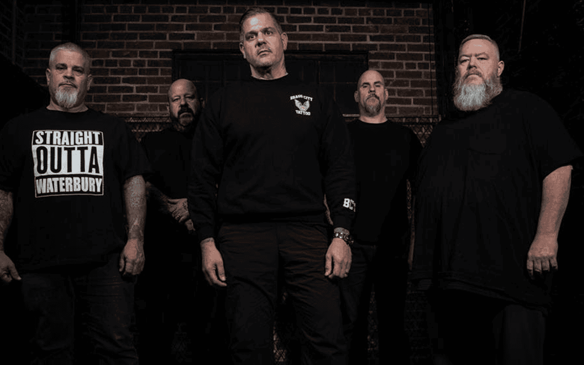 Five men in black shirts stand in a dark brick alley, posing as a rock/metal band promo photo.