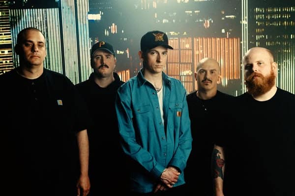 Five men pose as a band in a neon city backdrop; center wearer in a blue denim shirt and cap, others in black.