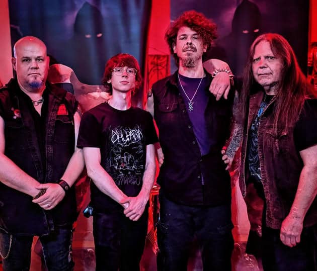 Four male rock musicians posing on a dimly lit stage with red lighting; from left, a bald man in a black vest, a young man with glasses, a tall man with curly hair, and a long‑haired man in a denim vest.