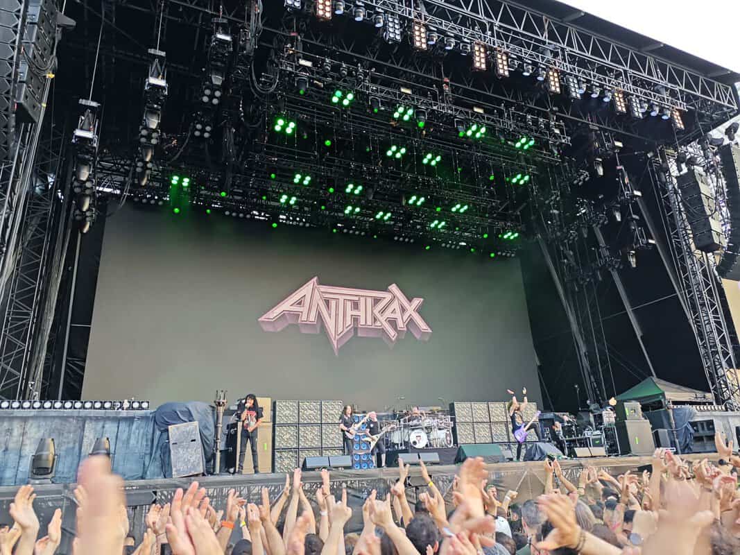 Concert stage with Anthrax logo on back wall, band performing while green lights shine and crowd raises hands.