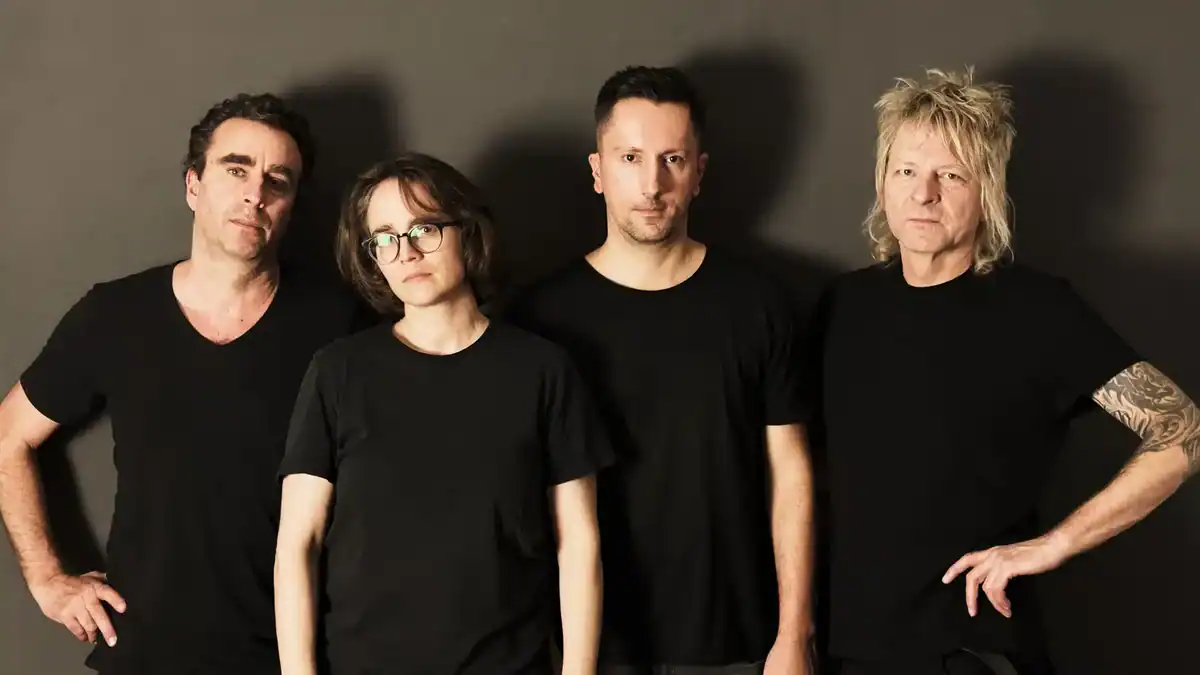Four adults wearing black T-shirts stand side by side against a dark backdrop, looking at the camera (band-style portrait).