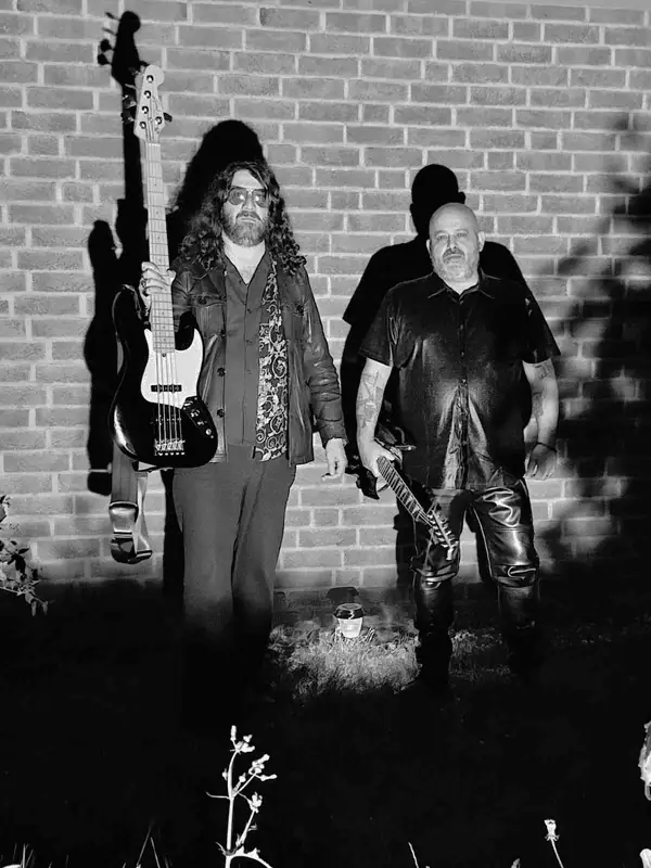 Two men stand against a brick wall at night, each holding a guitar: a bass guitar on the left and a smaller electric guitar on the right, all in a black‑and‑white photo with dramatic shadows.