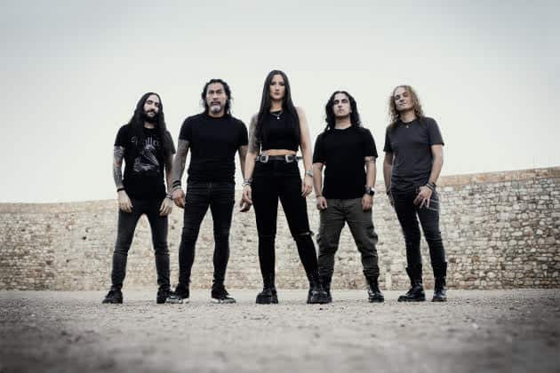 Five-member rock band posing together outdoors in dark clothing, standing in a line with a stone wall behind them.