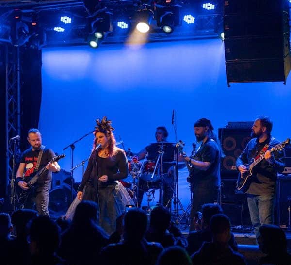 Band performing on a blue-lit stage: singer center with feathered headpiece, guitarists left and right, drummer in back, audience silhouettes in the foreground.