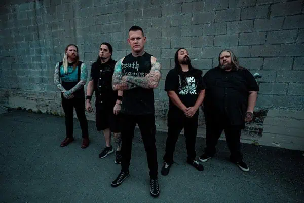 Five-member rock band posing in front of a gray brick wall; front man crosses arms in a sleeveless black shirt, tattoos visible, others in black attire standing behind.