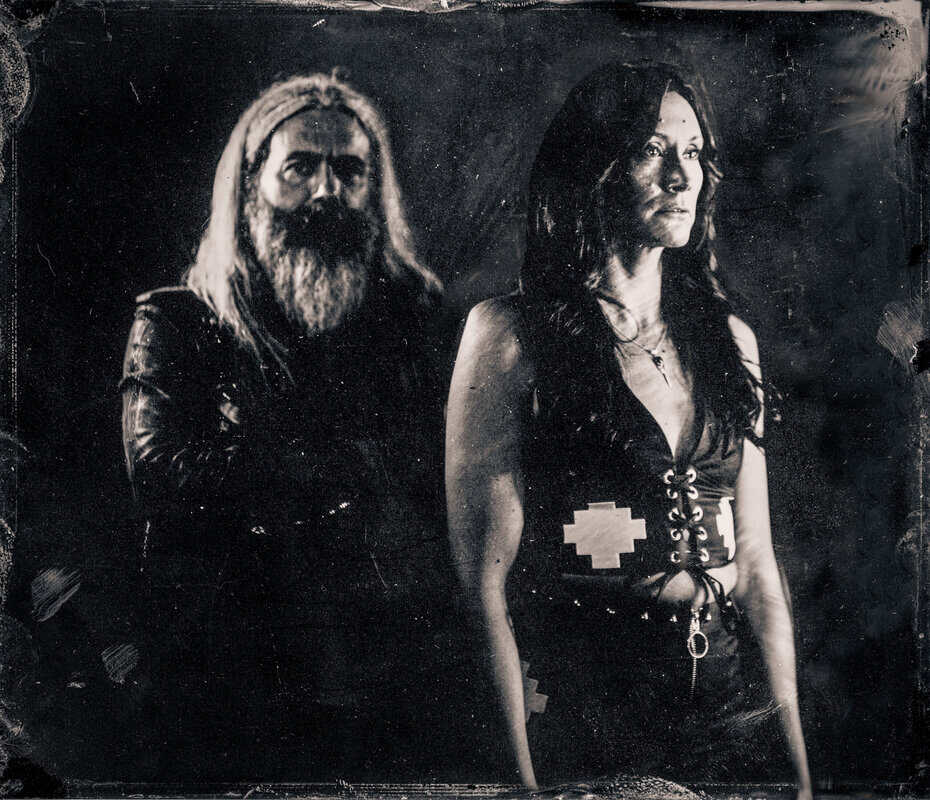 Black-and-white portrait of a bearded man with long hair standing behind a woman in a lace-up corset in dark clothing.