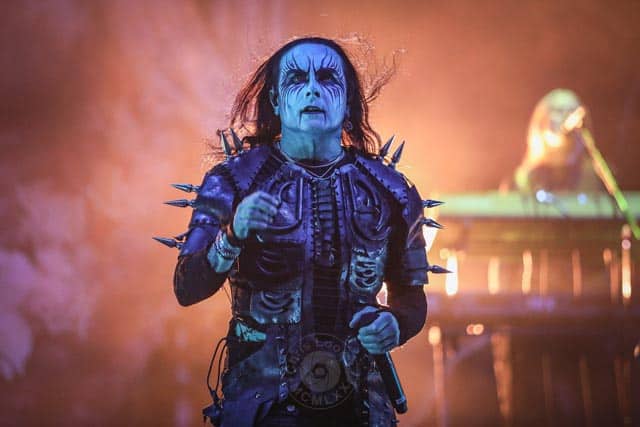 Male stage performer in blue makeup and spiked armor sings into a microphone amid orange stage lighting and smoky atmosphere when performing live.