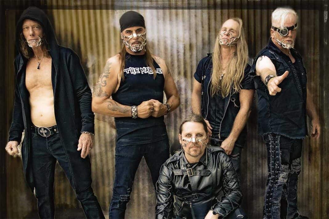 Five-member metal band in black leather and masked faces, posing against a rusted metal backdrop; one kneels front center while four stand behind.