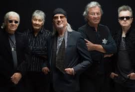 Five male musicians posing together for a band photo in black outfits against a dark backdrop.
