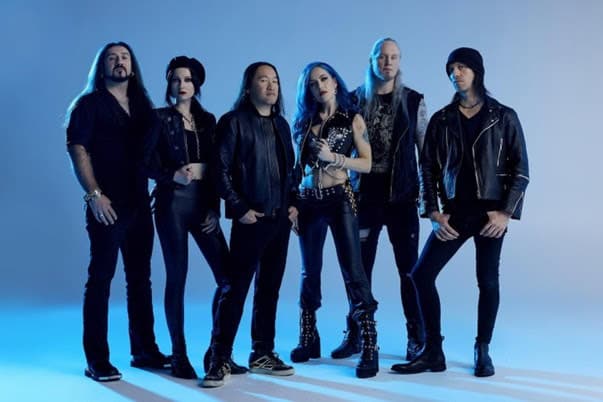 Seven-member rock band posing in a blue-toned studio, all wearing black leather jackets and dark outfits.