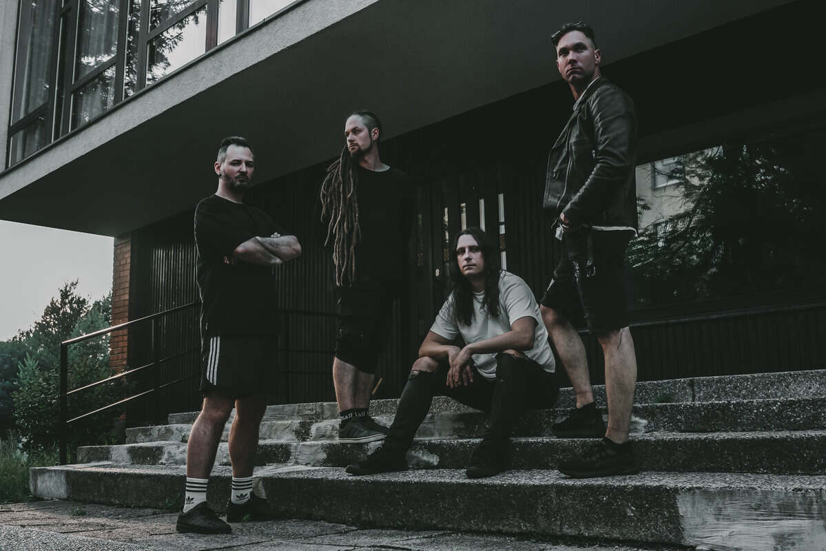 Four men posing on concrete stairs outside a modern building, casually dressed in dark clothes and boots, looking at the camera.