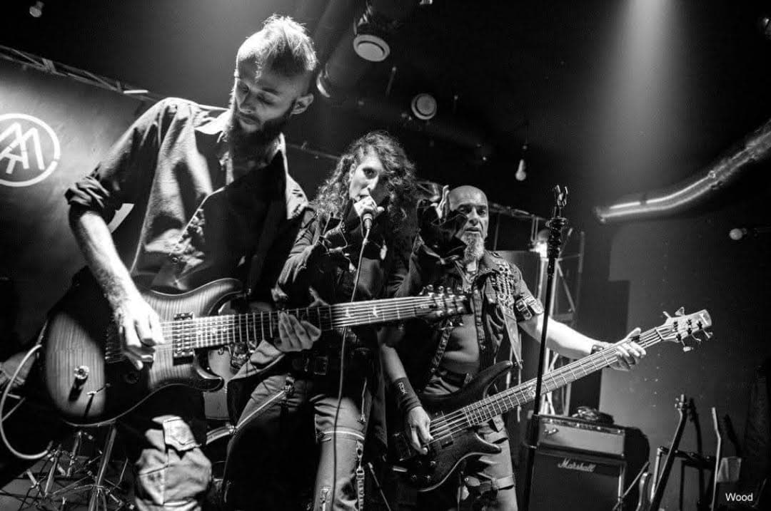 Three musicians perform on a dimly lit stage: a guitarist left, a vocalist center with a microphone, and a bassist right.