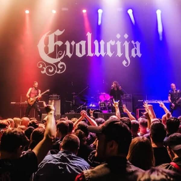 Live rock band on stage with a large Evolucija banner, colorful lights, and a crowd raising their hands in front of the stage.