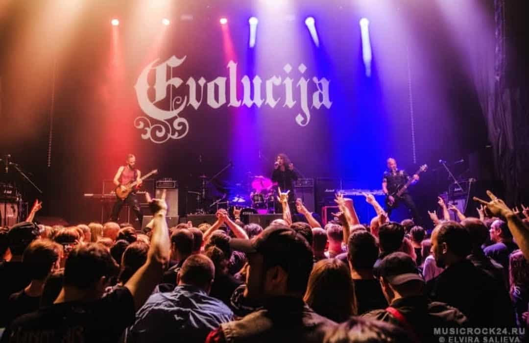 Live rock band on stage with a large Evolucija banner, colorful lights, and a crowd raising their hands in front of the stage.