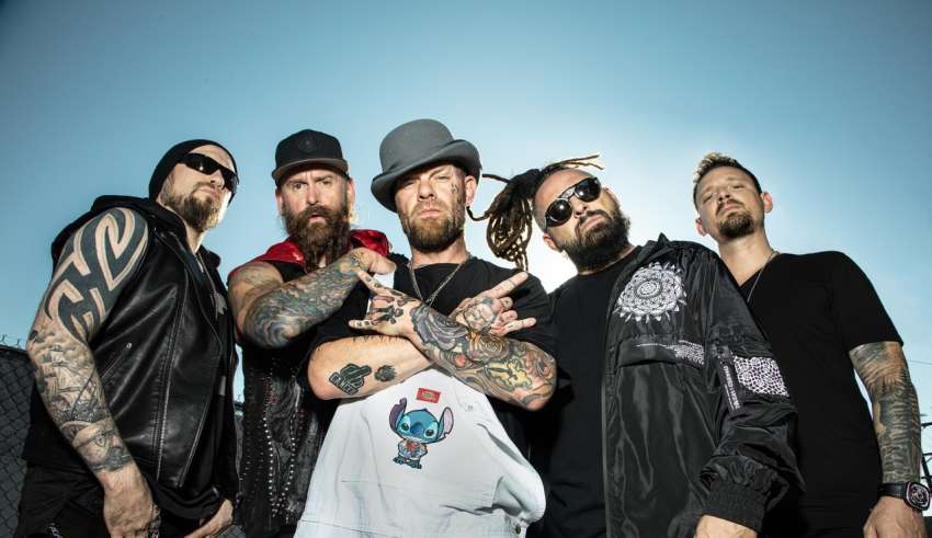 Six tattooed men in dark clothing pose together outdoors against a blue sky in a promo-style shot.