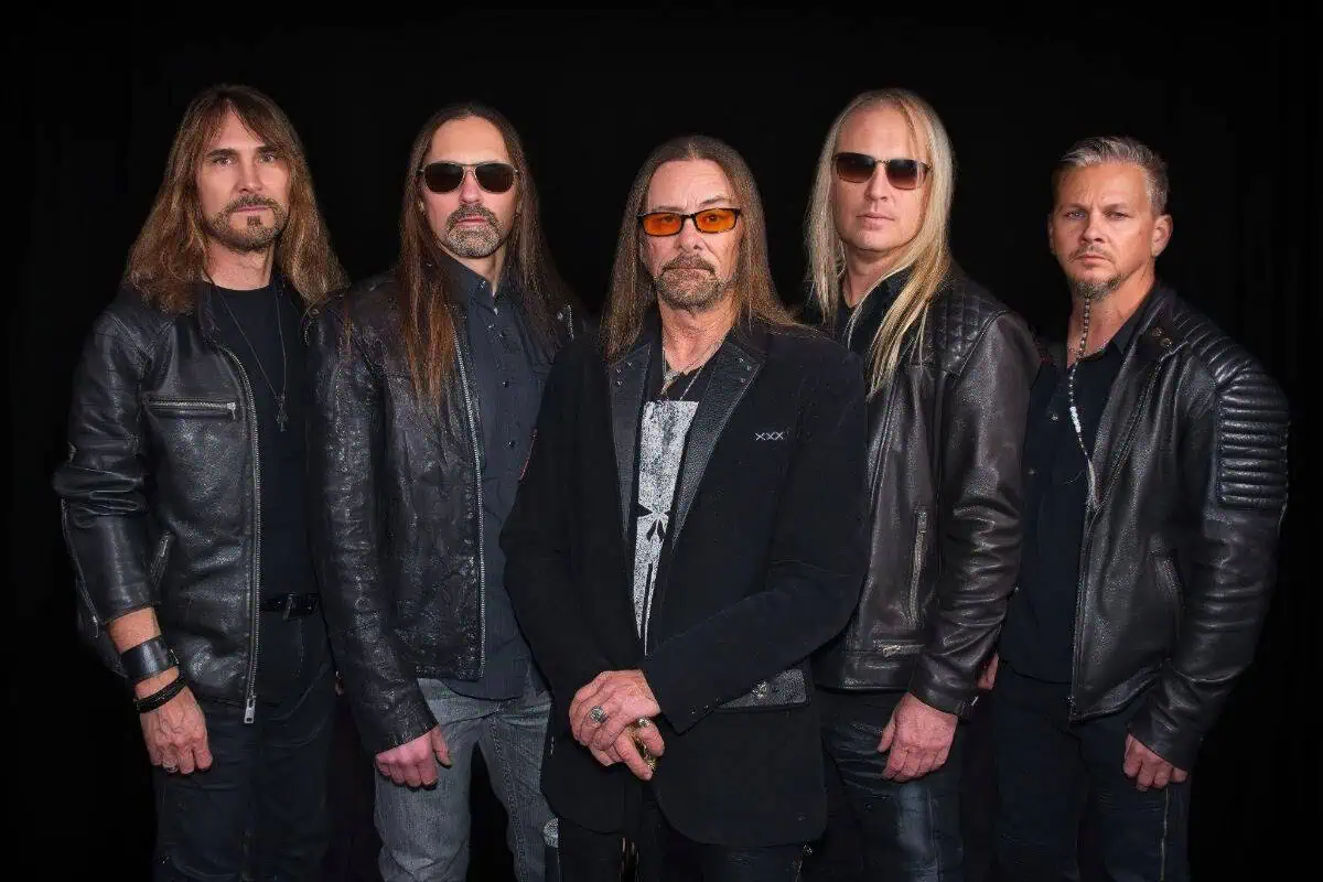 Five male rock musicians in black leather jackets posing against a dark backdrop.