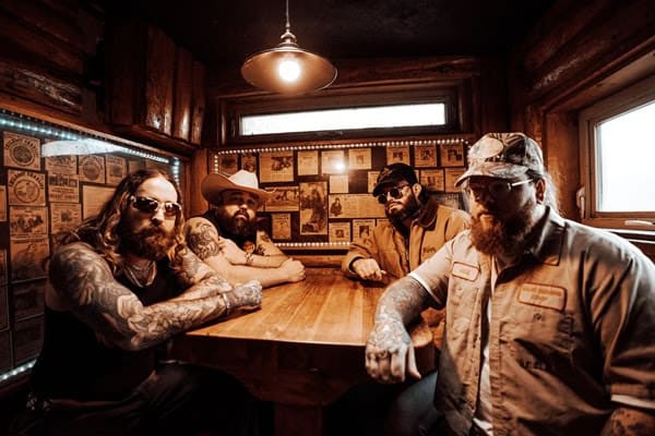 Four men with beards and tattoos sit around a wooden bar in a dim rustic room, posing for a photo.