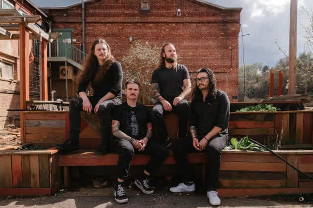 Four men with long hair and beards pose on wooden benches outdoors in front of a brick building.