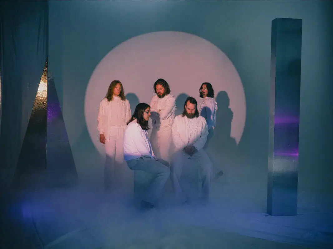 Six people in white robes in a foggy studio, seated and standing, with a large circular light behind them.