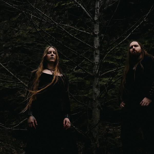 Two people in dark robes stand in a dim forest with bare branches, looking toward the camera.