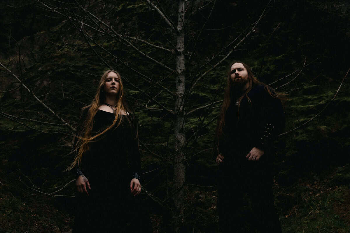 Two people in dark robes stand in a dim forest with bare branches, looking toward the camera.