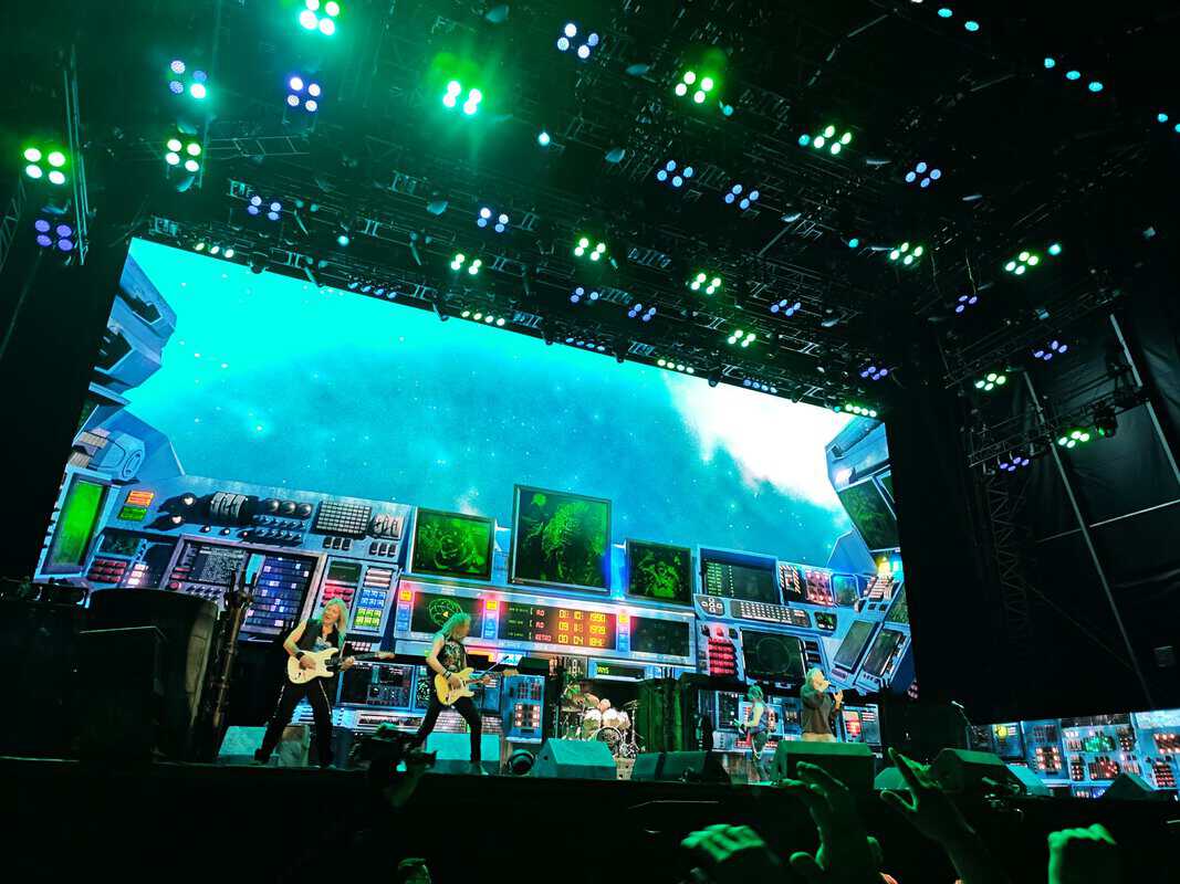 Rock band performing on a neon-lit, sci-fi inspired stage with synthesizer panels and bright green and blue lights from above and around.