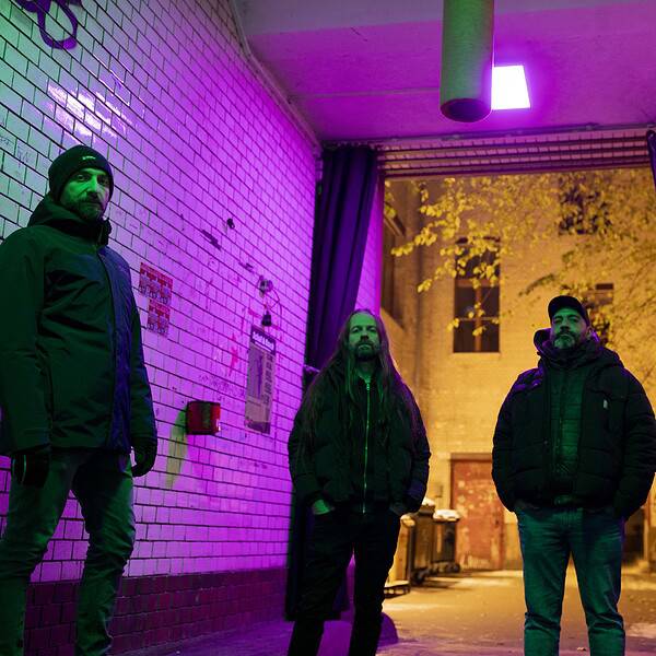 Four men stand in a narrow brick alley bathed in purple and green neon light, wearing winter jackets and looking at the camera.