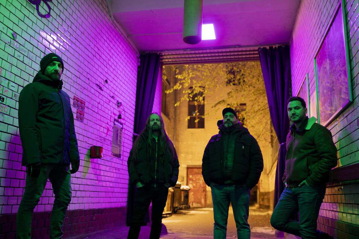 Four men stand in a narrow brick alley bathed in purple and green neon light, wearing winter jackets and looking at the camera.
