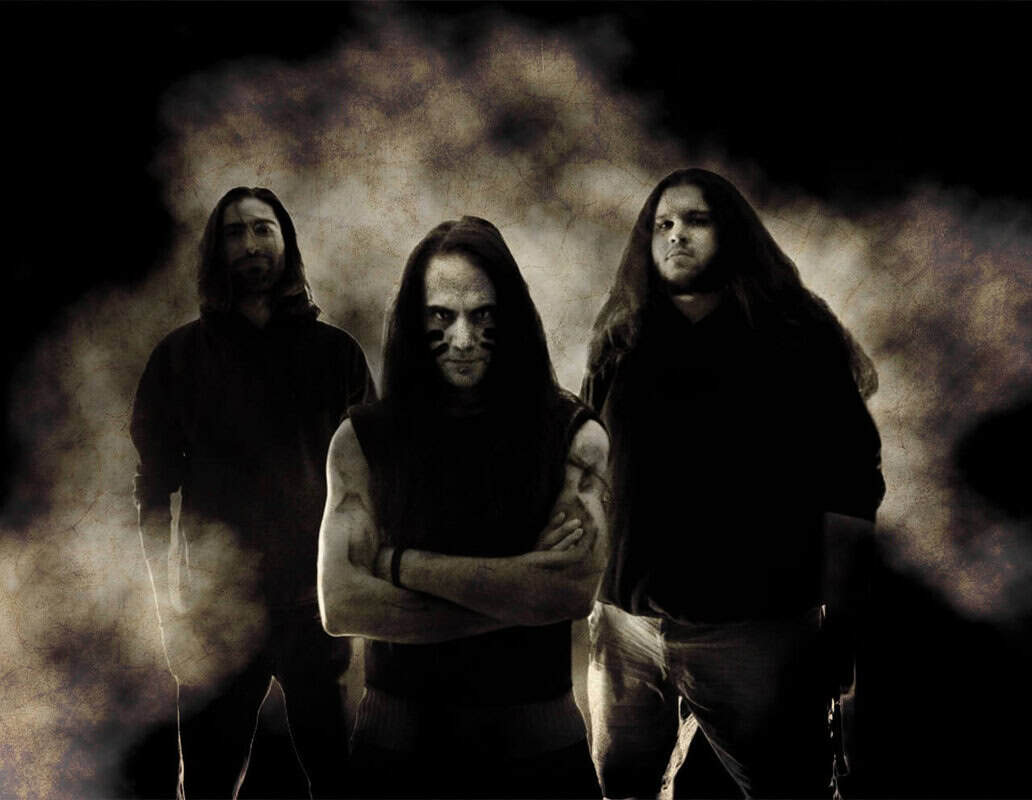 Three men in a moody, smoky photo; central figure crosses his arms, flanked by two others in shadowy lighting.