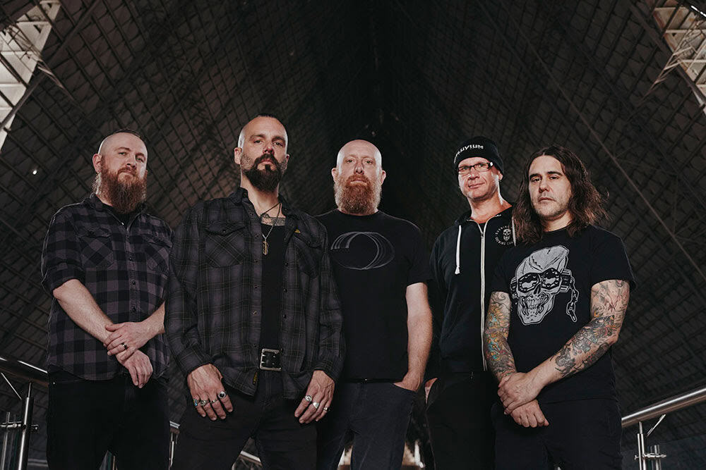 Five male rock or metal musicians pose together in a dim, industrial setting with a lattice ceiling behind them.