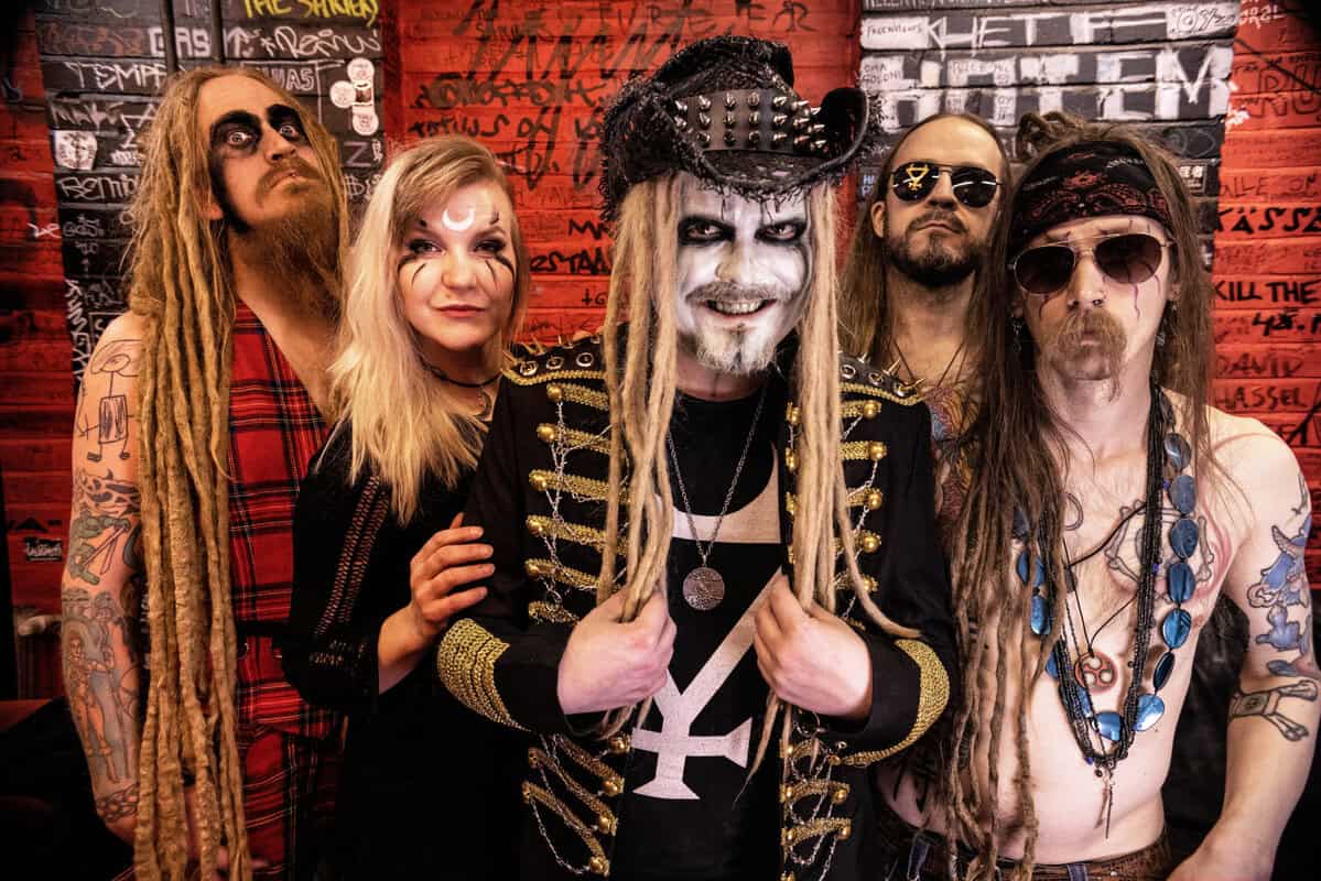 Group of five people in heavy-metal costumes posing against a graffiti-covered red wall, all with distinctive makeup and accessories.