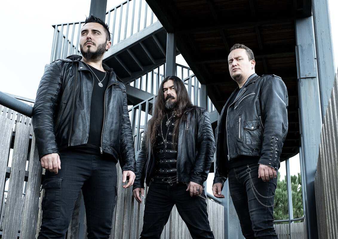 Three men in black leather jackets pose on an industrial stairway, looking serious and ready to perform.