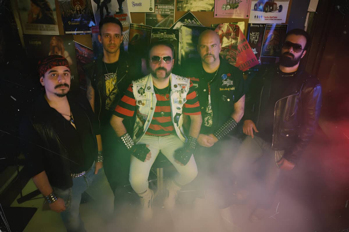 Six men in punk/rock outfits pose in a dim, poster-covered room with tinted green and red smoke adding a moody atmosphere.