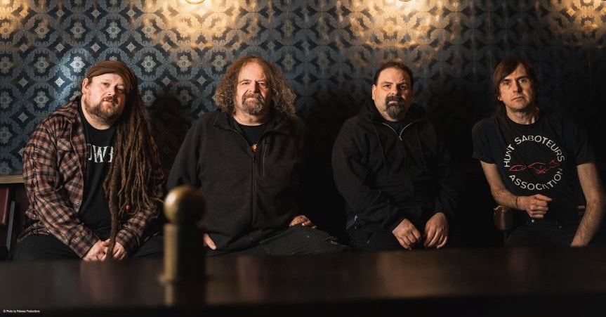 Four men with long hair sit at a dark bar against a vintage patterned wall, posing as a rock band.