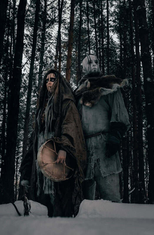 Two costumed figures in a snow-covered forest, one with a white mask and black streaks, the other with a darker mask, carrying a drum and wearing fur robes.