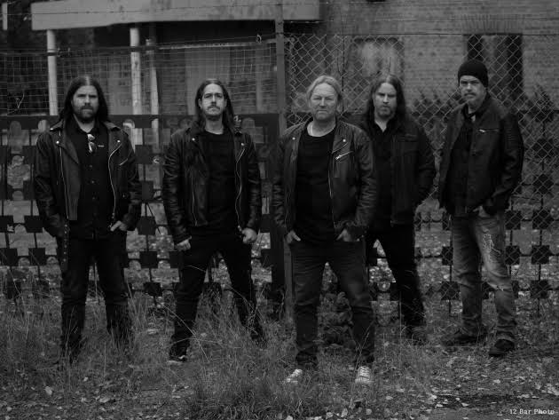 Five men in black leather jackets stand side by side in a gritty outdoor area with a chain-link fence behind them, suggesting a rock band photo.