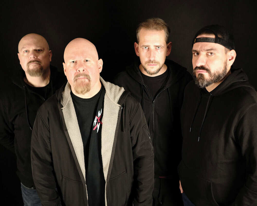Four men in dark hoodies posing against a black background, serious expressions; one in a coat with a light fleece lining, another wearing a backward cap (group photo).