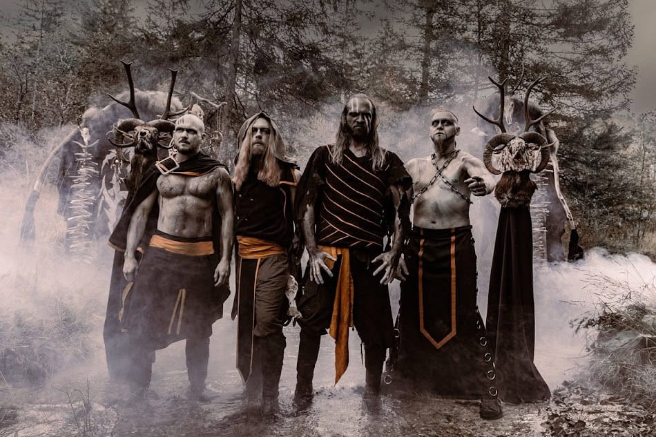 Six men in black and orange fantasy costumes pose in a smoky forest clearing, wearing armor-like belts and cloaks.