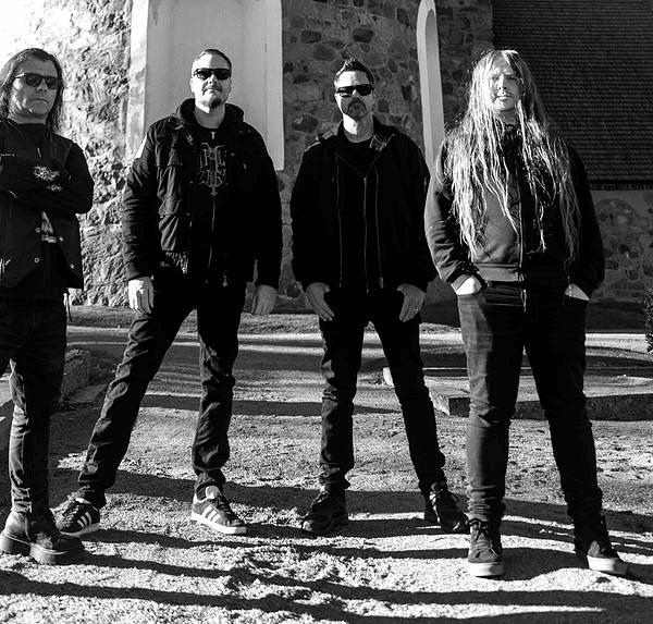 Four men in dark clothing and sunglasses pose on stone steps outside a brick building, forming a band photo.