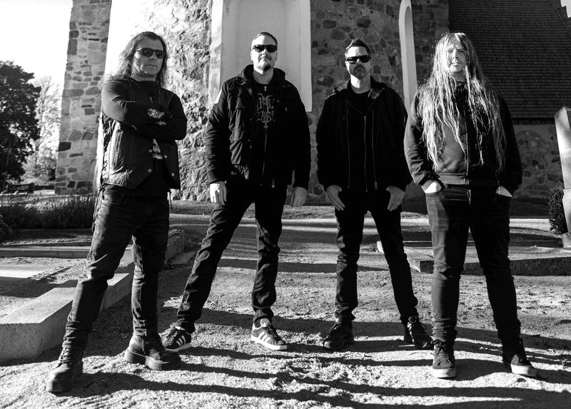 Four men in dark clothing and sunglasses pose on stone steps outside a brick building, forming a band photo.