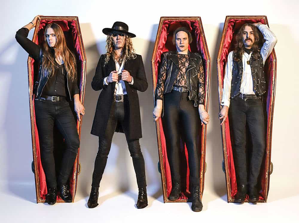 Four rock musicians pose standing inside open coffins with red interiors, wearing black leather and dark clothing.