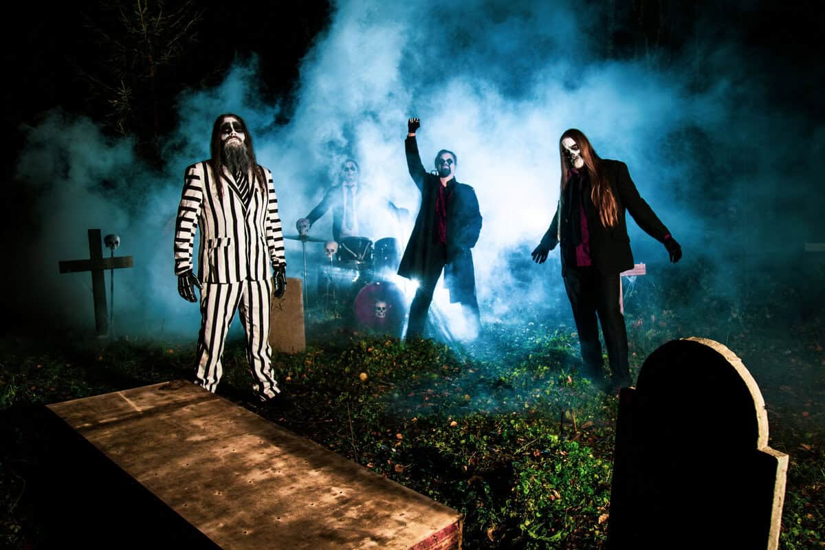 Gothic rock band performs outdoors at night, blue fog surrounding members with skull makeup and dark costumes.