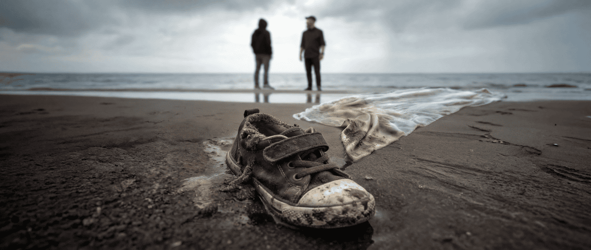 Worn, muddy sneaker and a torn cloth lying on a damp beach; two figures stand near the water in the distance.