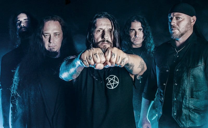 Five men in a metal band pose in a dark studio; center figure extends fists toward the camera, tattoos visible on his hands.
