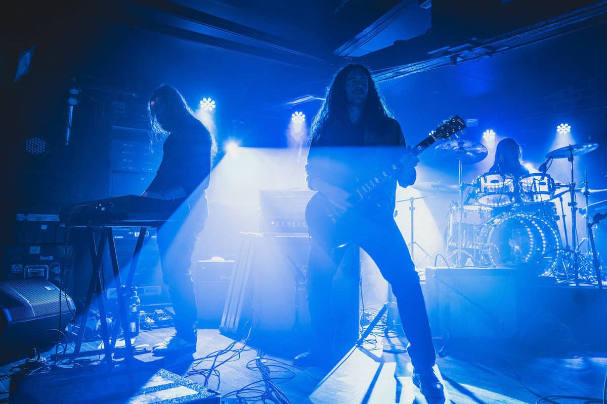 Three musicians on a blue-lit stage: a guitarist center, a keyboardist left, and a drummer at the back with drums visible.