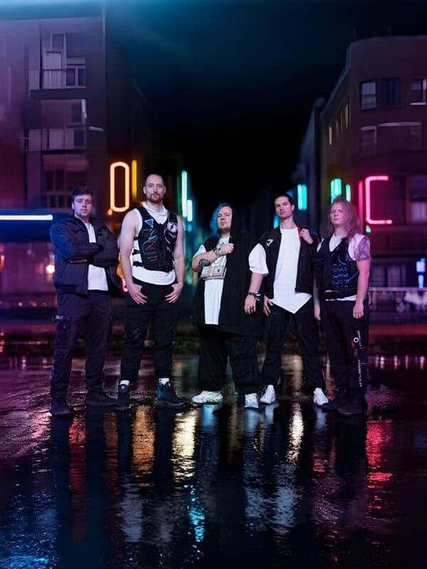 Five men stand together on a neon-lit city street at night, posing as a band with reflections shimmering on the wet pavement.